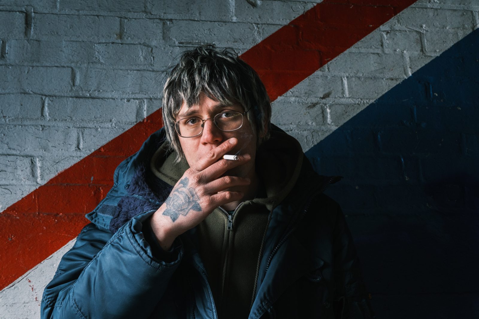 Mendez, wearing a blue puffer coat stands in front of a red, blue and white stripped wall smoking a cigarette.